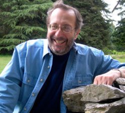 Smiling man with blue denim shirt sitting outside.